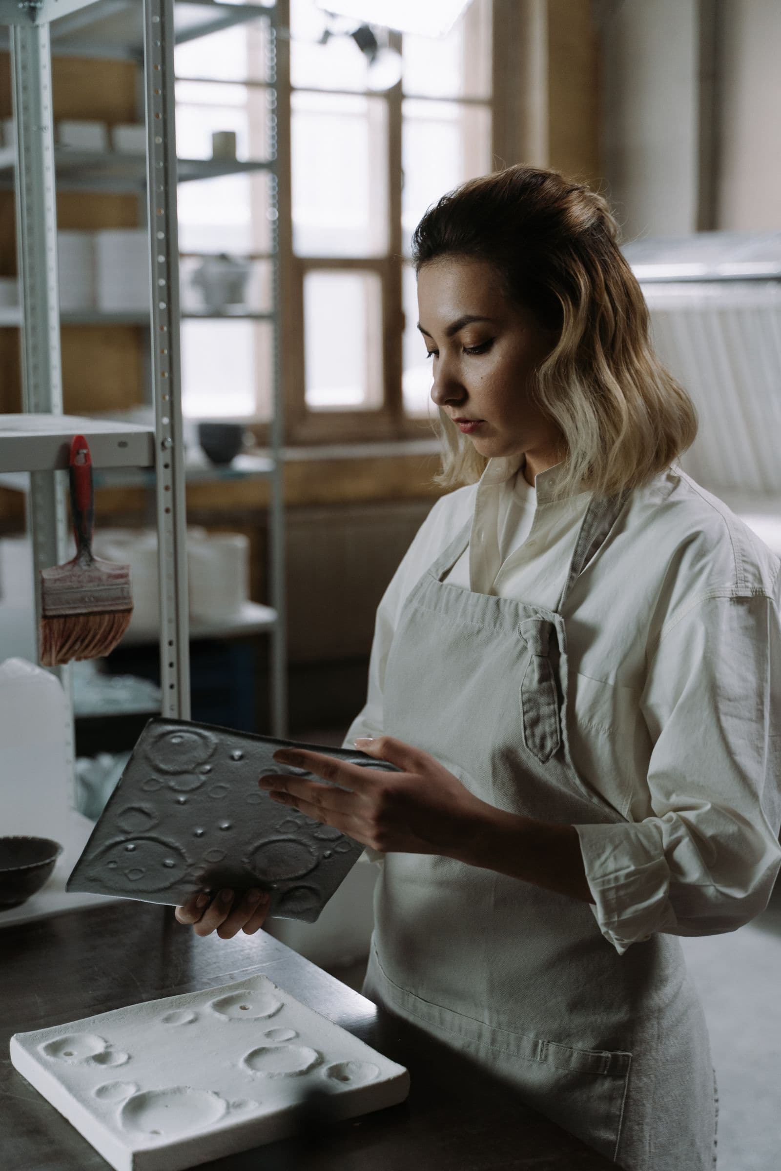 A maker in a workshop apron examining a cast mould by window light