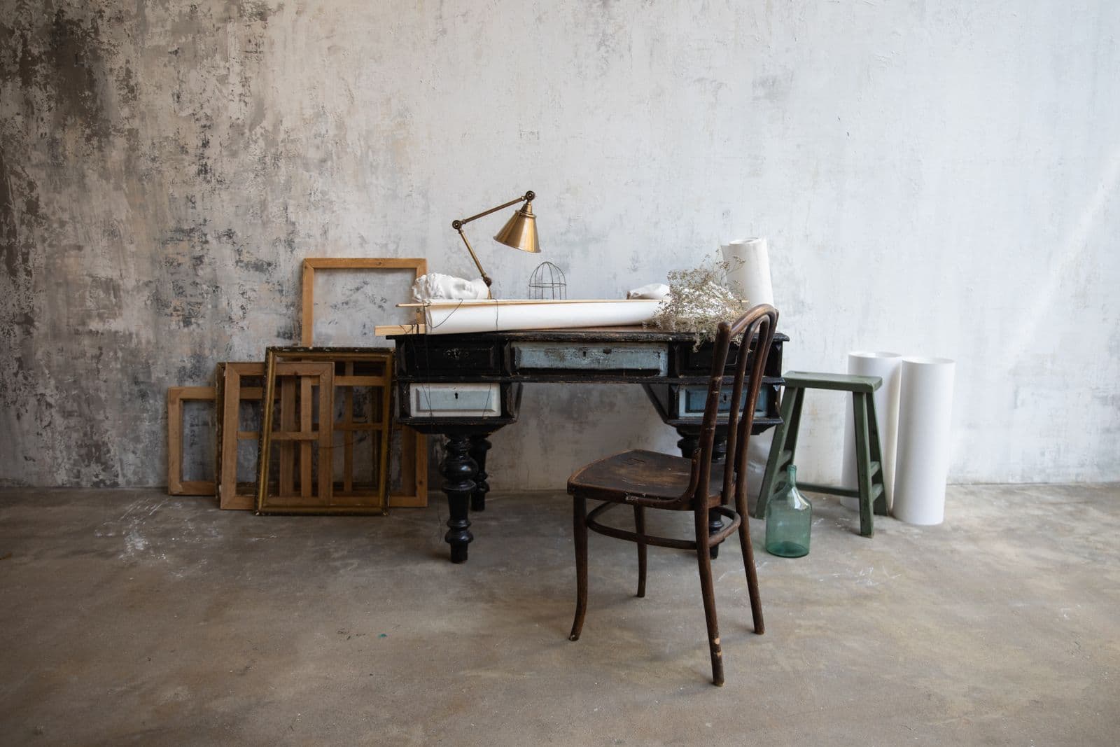 An empty workroom — desk, lamp, wooden chair and stacked frames against a plaster wall