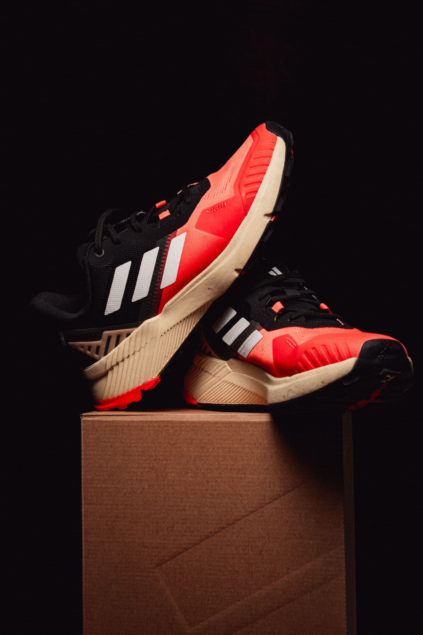 Red-and-black trail sneakers posed on a kraft box against a black backdrop
